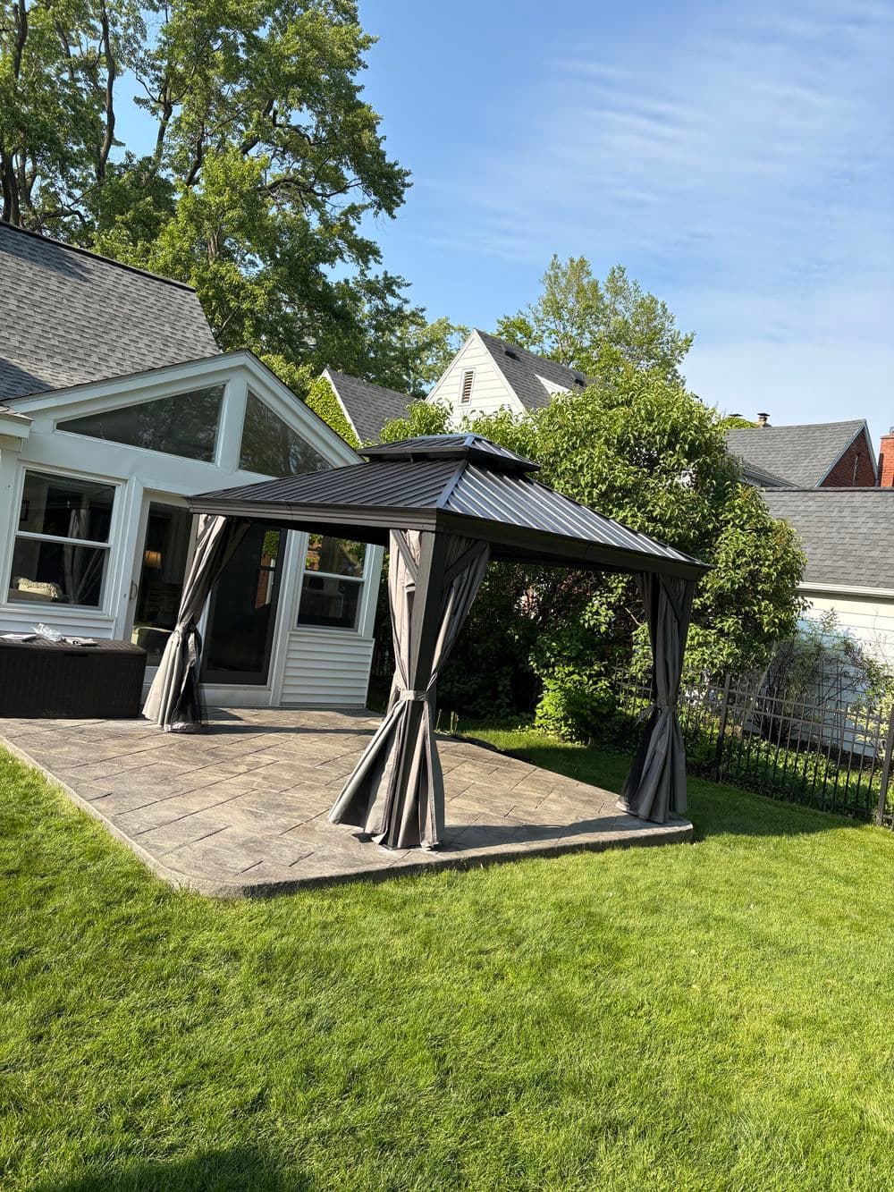 Modern backyard gazebo with dark curtains on a lawn, surrounded by greenery and houses.
