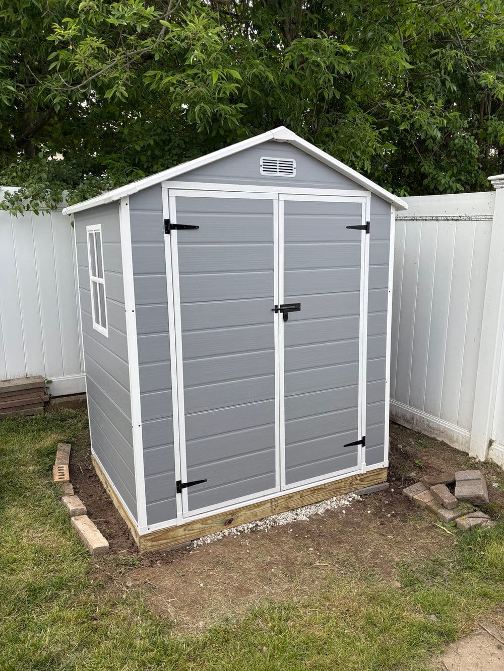 Gray outdoor shed with double doors and window, surrounded by grass and a fence.