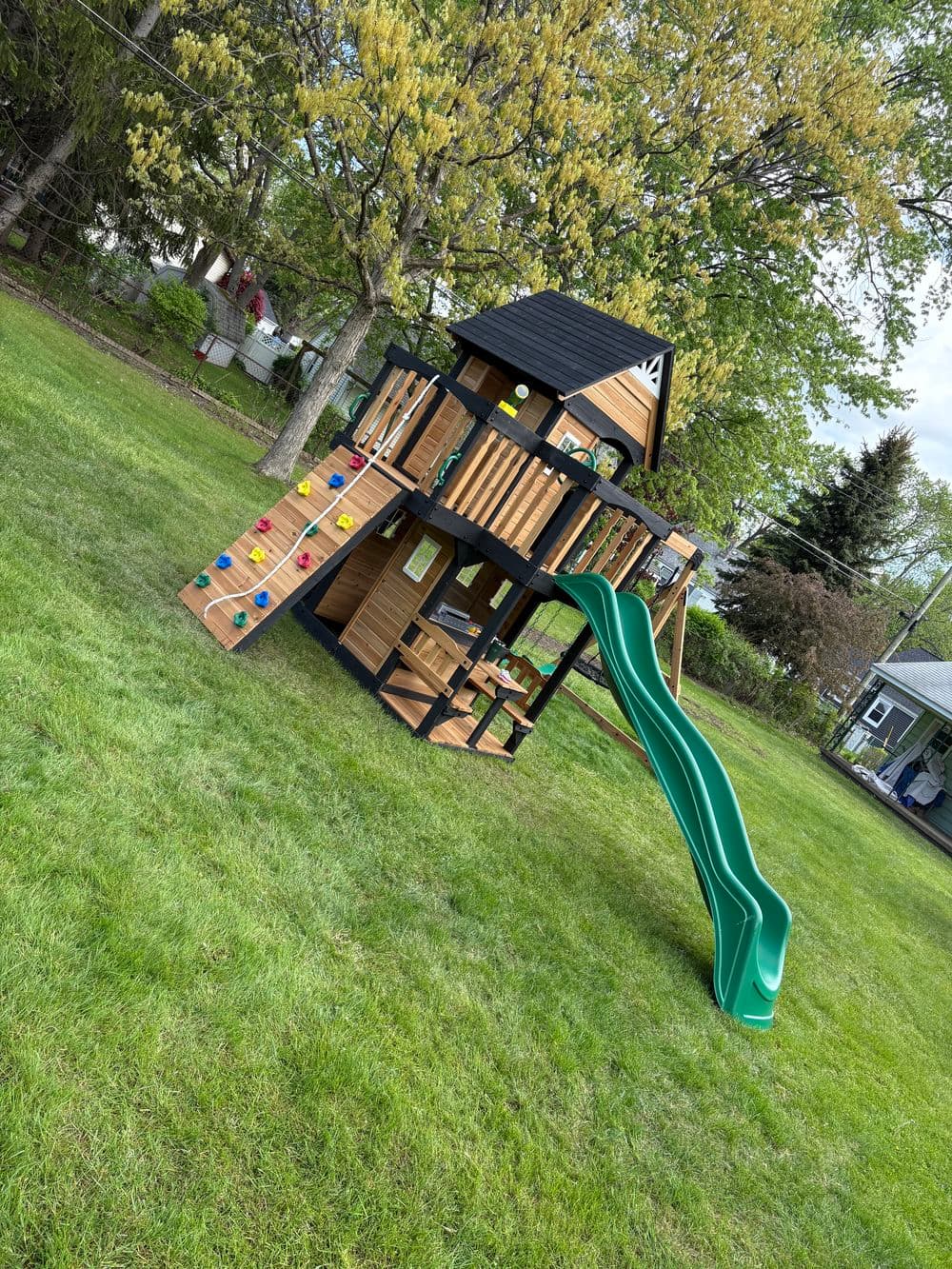 Wooden playset with climbing wall and slide in a green backyard. Ideal for children's outdoor fun.