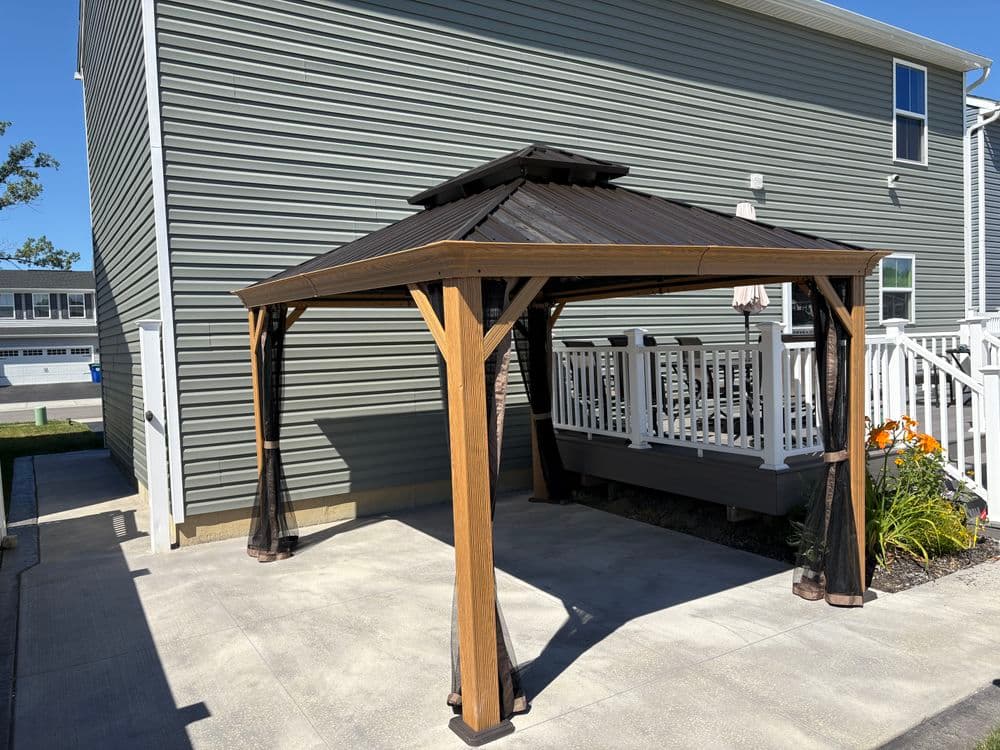 Wooden gazebo with a metal roof set on a patio beside a house and garden.