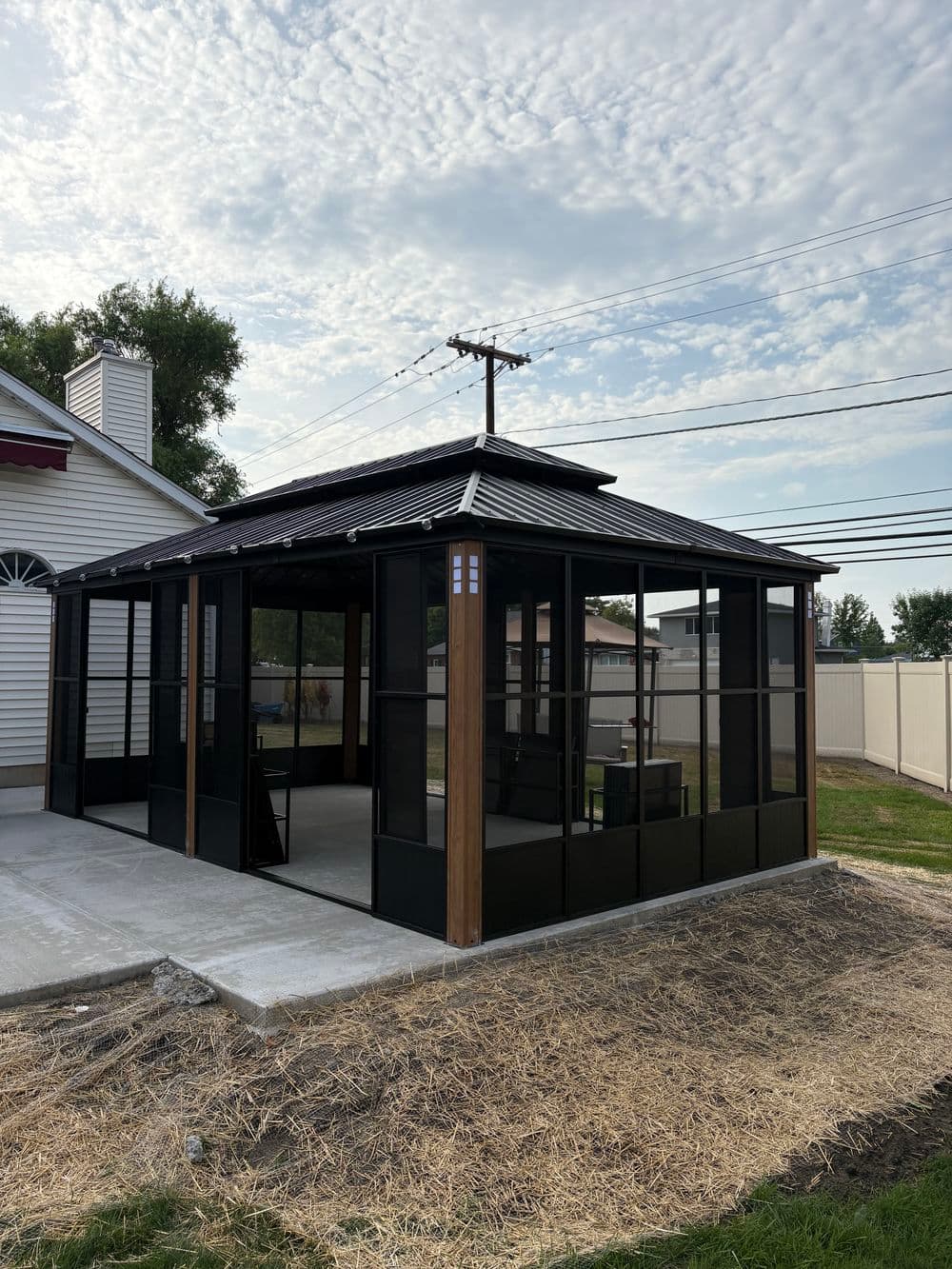 Modern black screened-in gazebo with a patio, surrounded by grass and power lines.